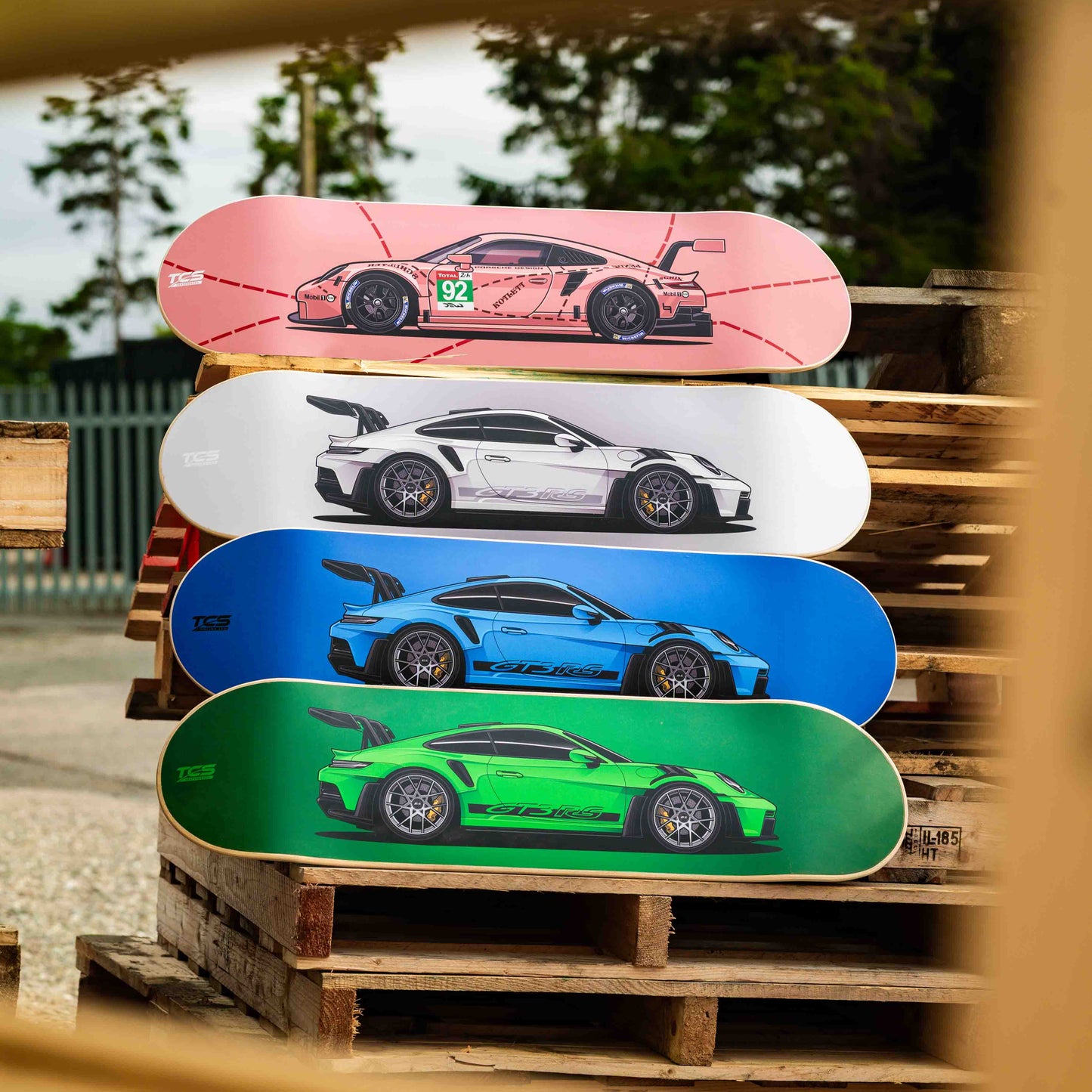 Stack of skateboard decks with car designs on a wooden pallet outdoors.