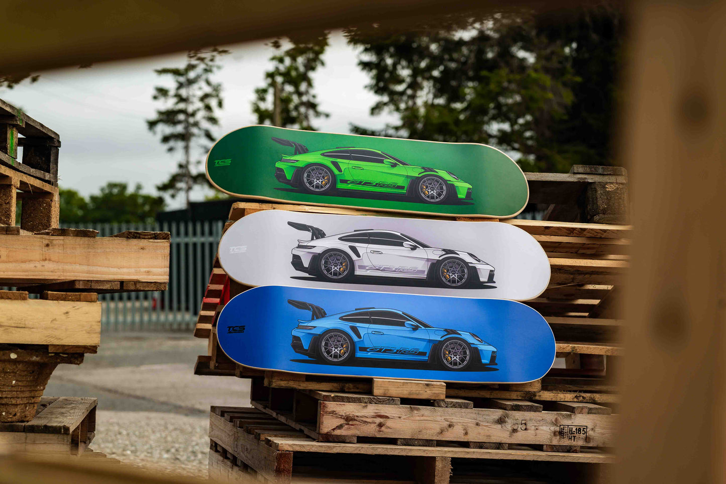 Three skateboard decks with car designs stacked on a wooden pallet outdoors.