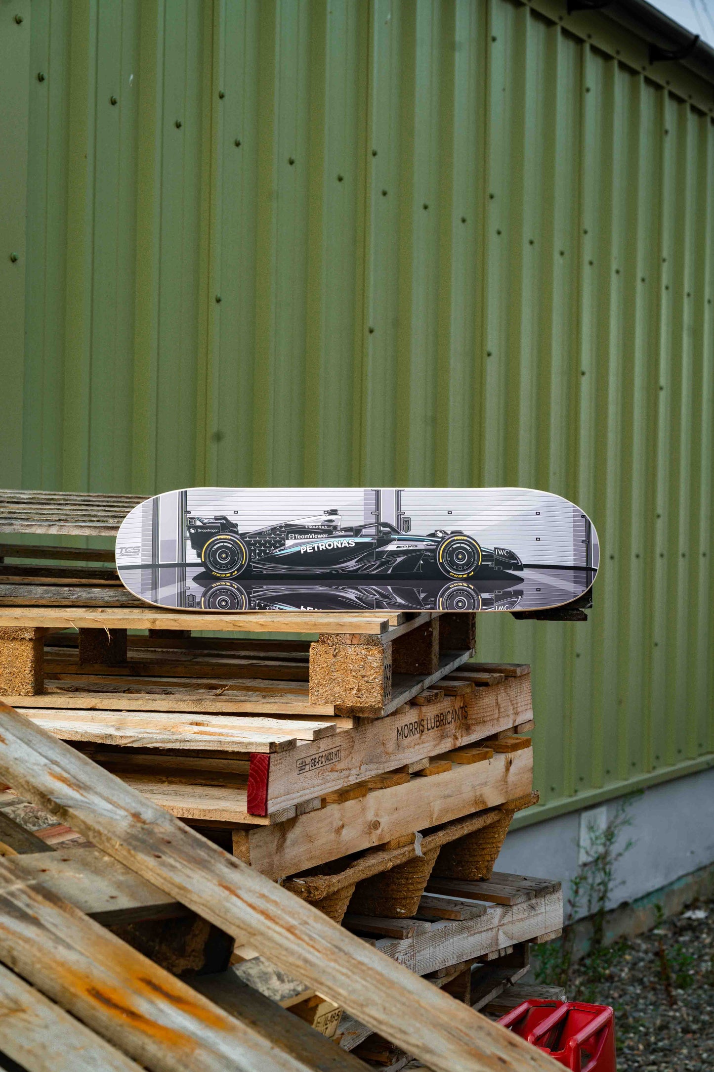 Skateboard with a Formula 1 car design on a wooden pallet against a green corrugated metal wall.