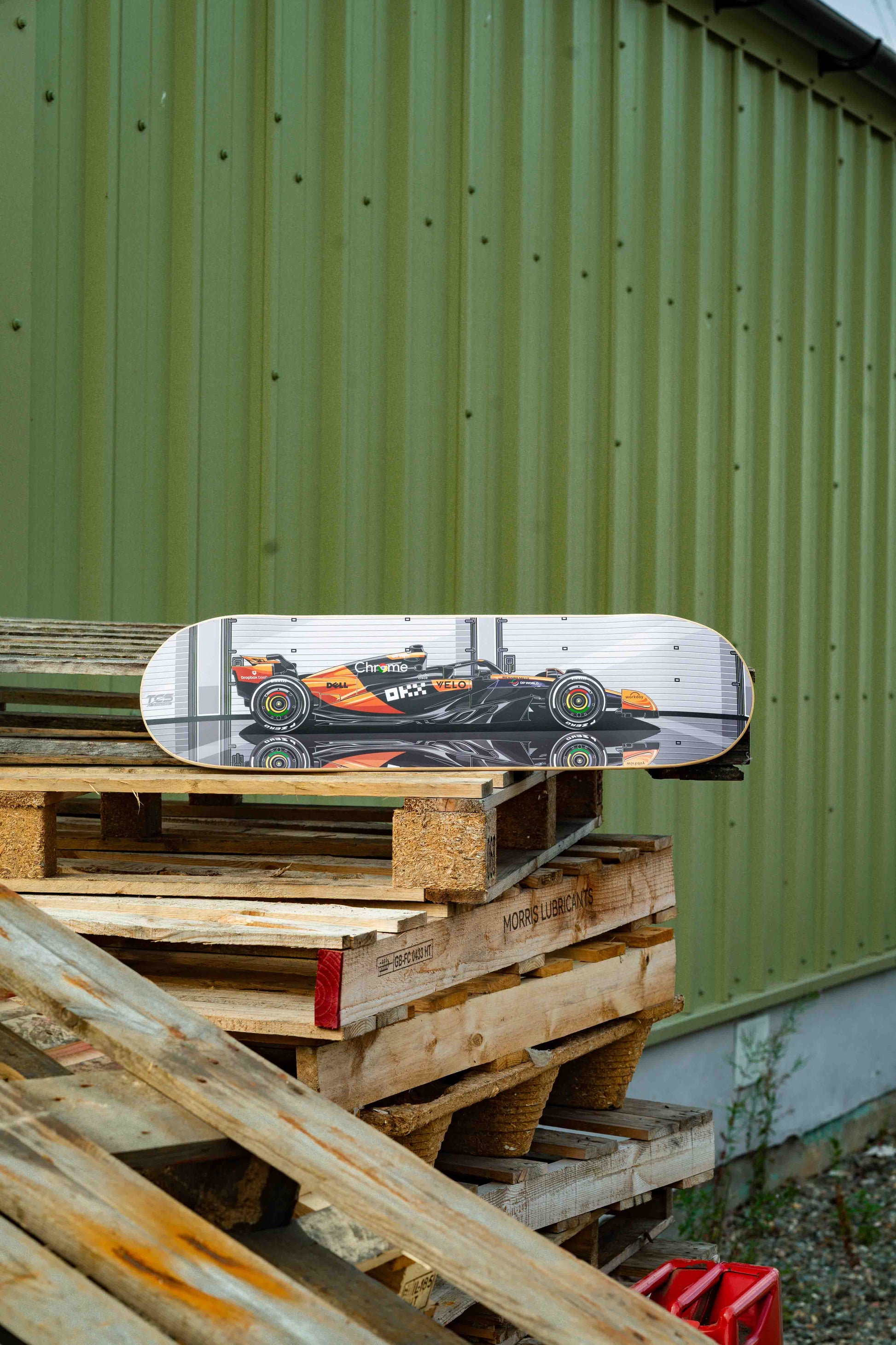 Skateboard with a Formula 1 car design on a stack of wooden pallets against a green wall.