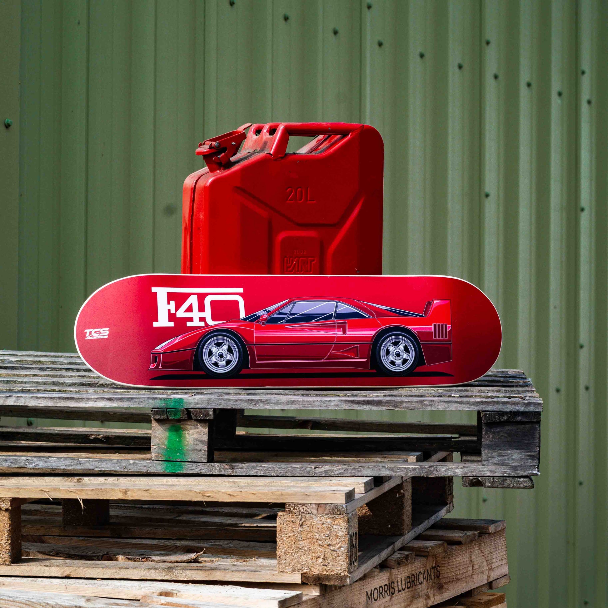 Red skateboard with a car design on a wooden pallet against a green corrugated metal wall.