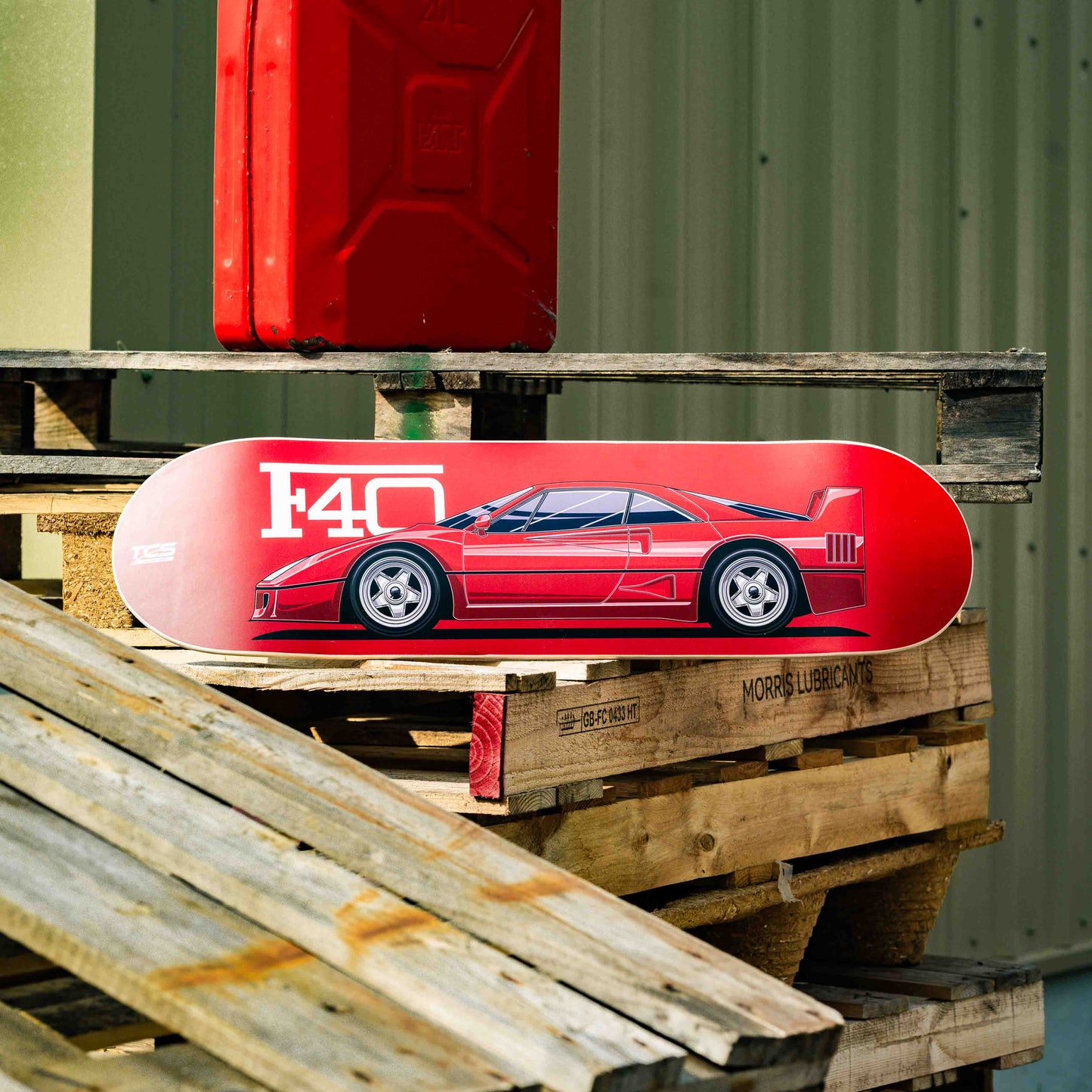 Red skateboard deck with a car design on a wooden pallet against a green metal wall.