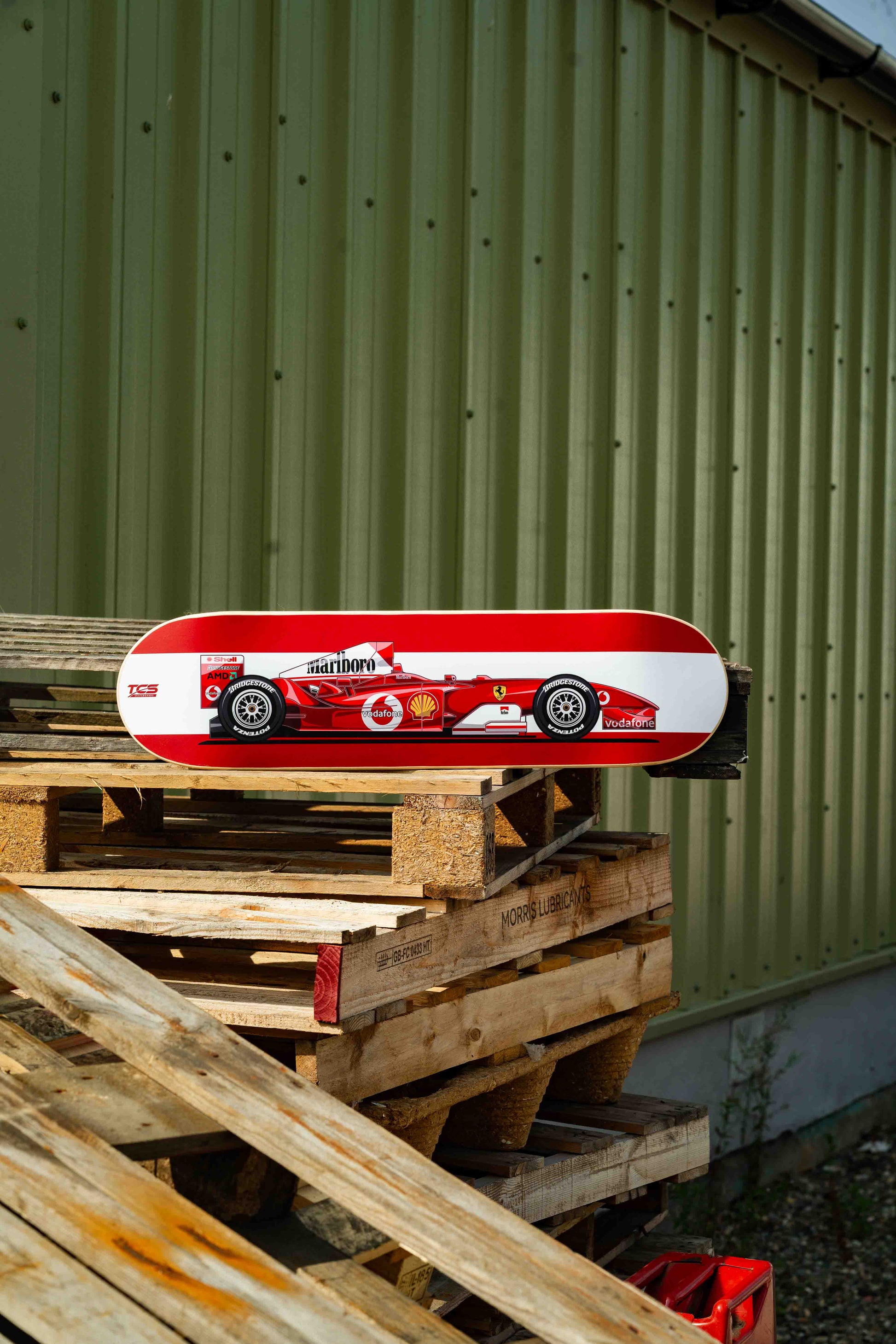 Red and white skateboard with a racing car design on wooden pallets against a green wall.