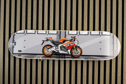 A skateboard deck featuring a graphic print of a Honda Repsol racing motorcycle, displayed flat against a vertical wood slat wall.