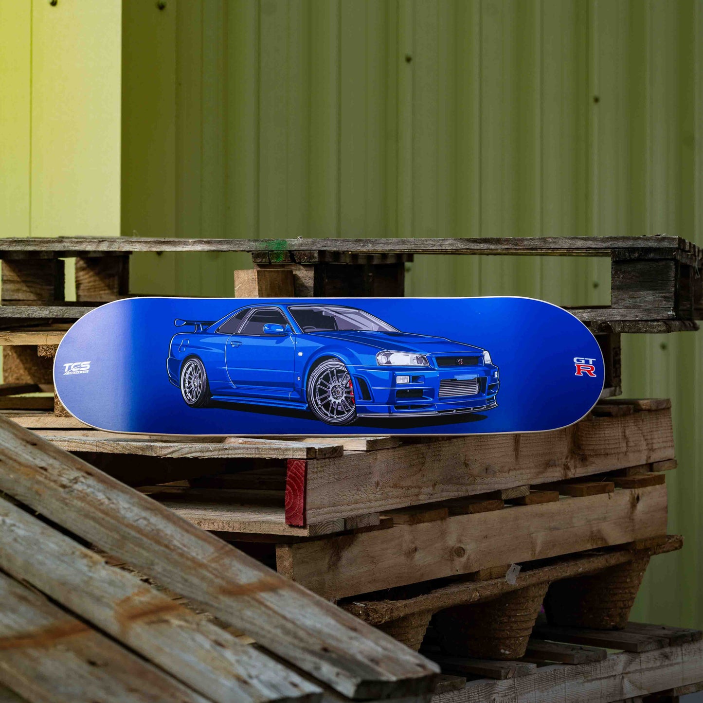 Blue car-shaped object on wooden pallets with a green corrugated metal wall in the background