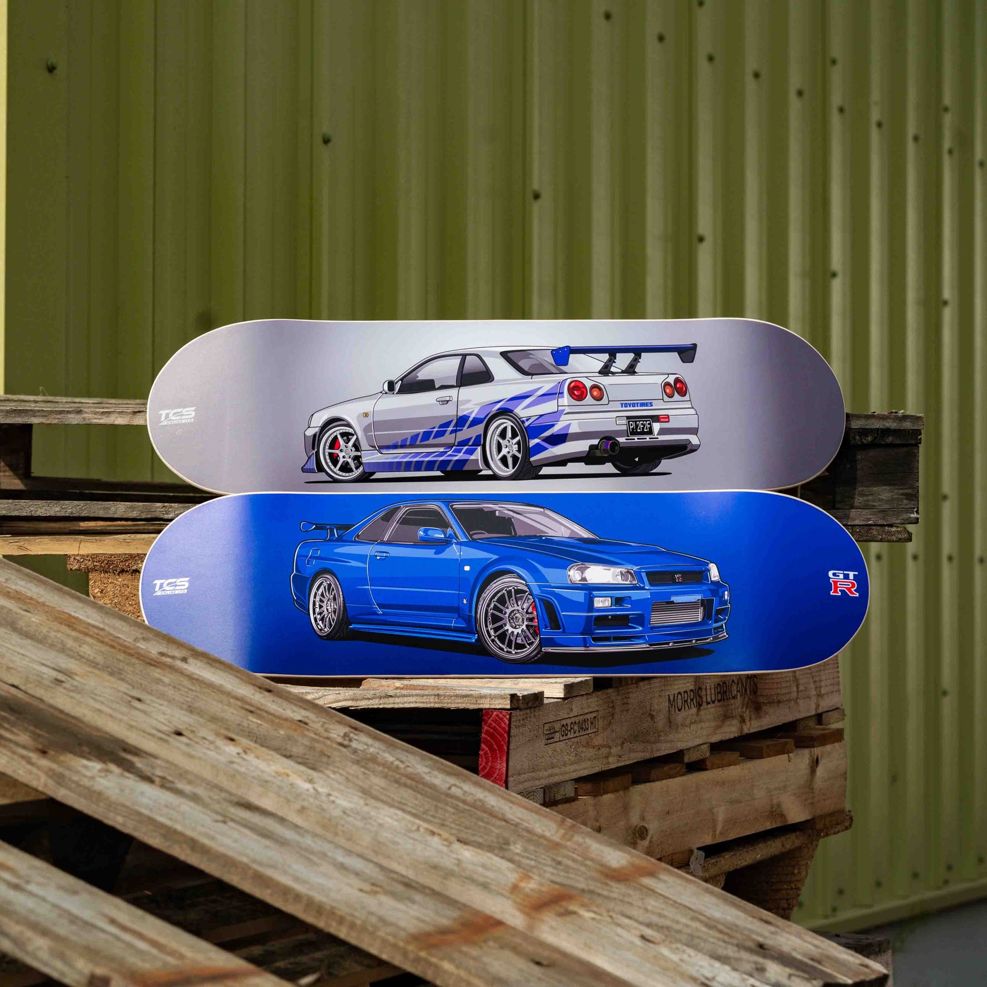 Two skateboard decks with car designs on a wooden pallet against a green corrugated metal wall.