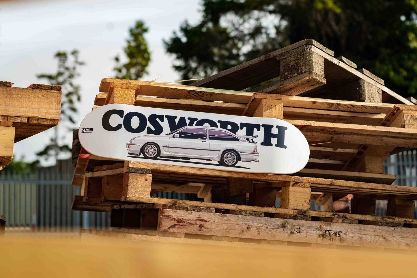 Cosworth logo on a skateboard deck placed on wooden pallets outdoors