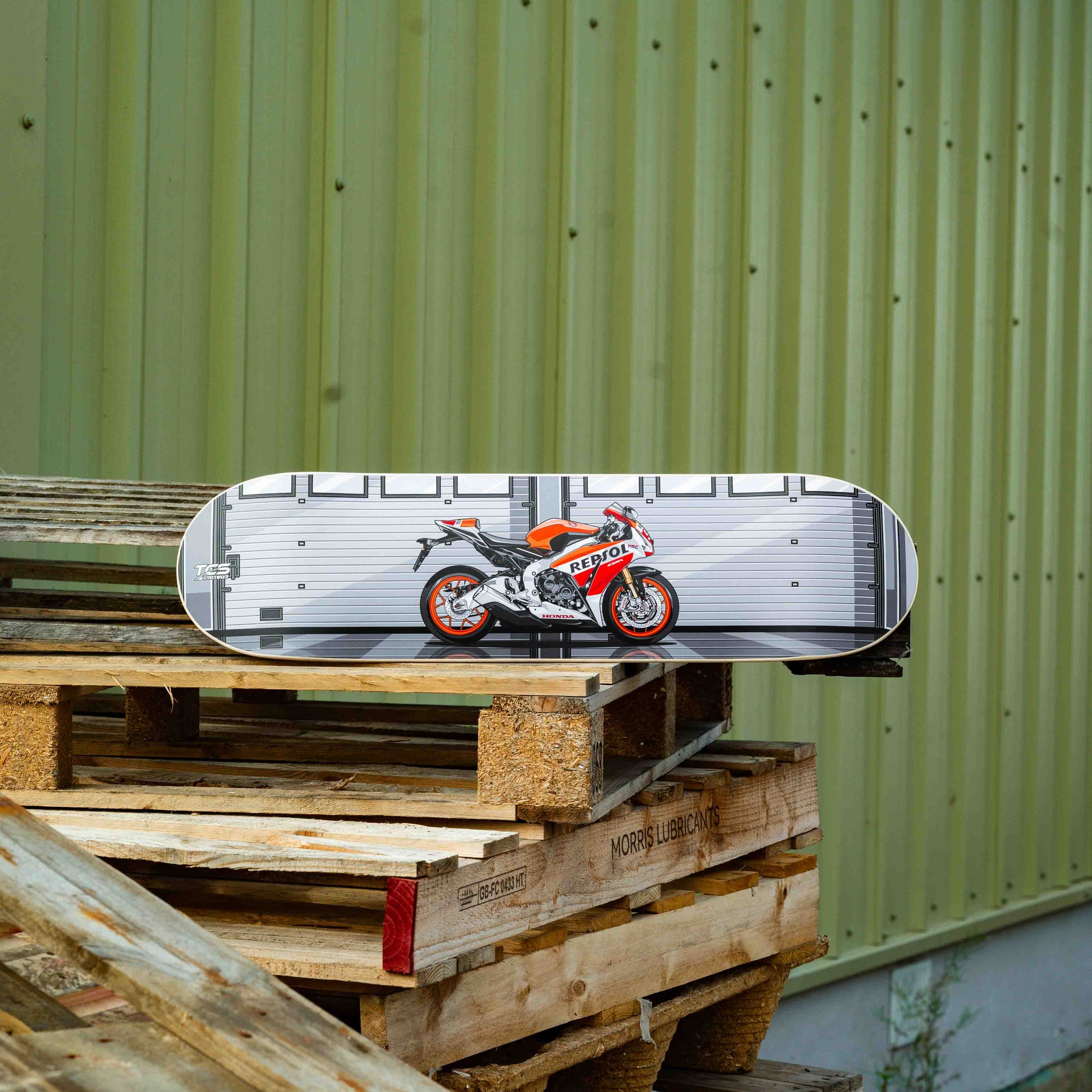 Skateboard with a motorcycle design on a wooden pallet against a green wall.