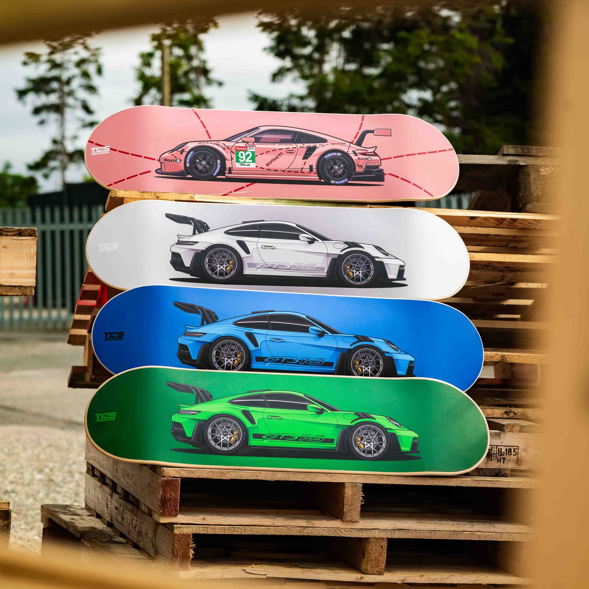 Stack of skateboard decks with car designs on a wooden pallet outdoors.