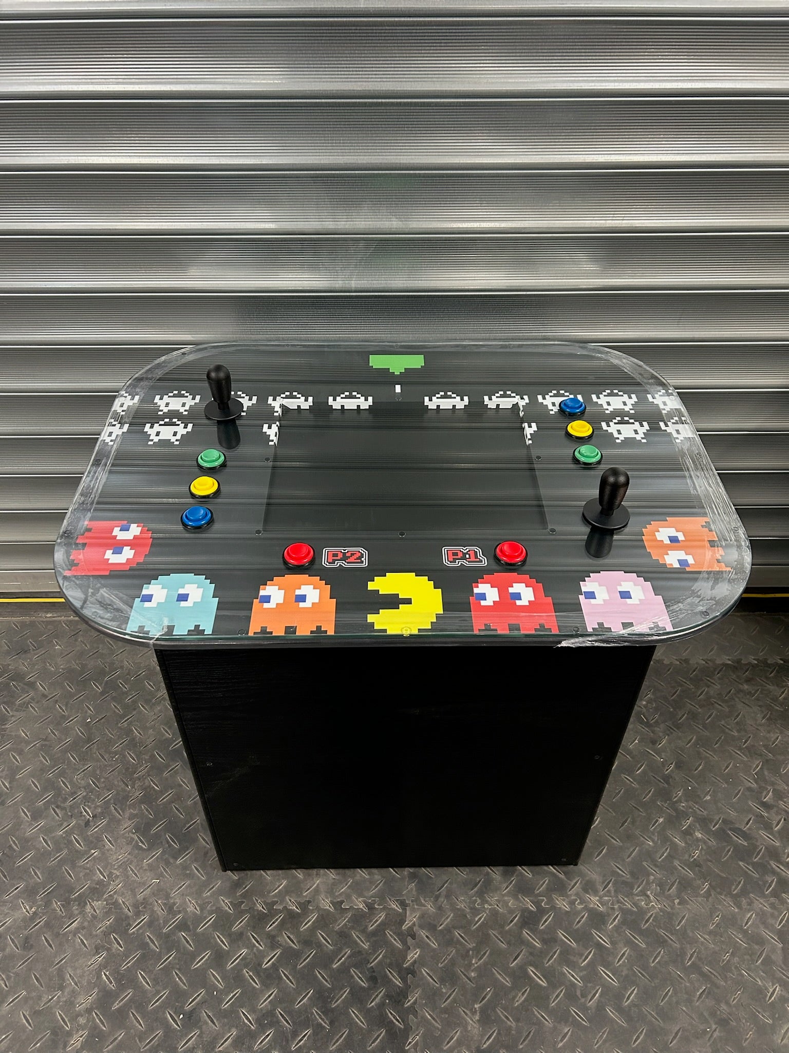A retro arcade cocktail table with a glass top featuring colorful arcade game icons, built-in speakers, and adjustable feet, set against a metallic background.