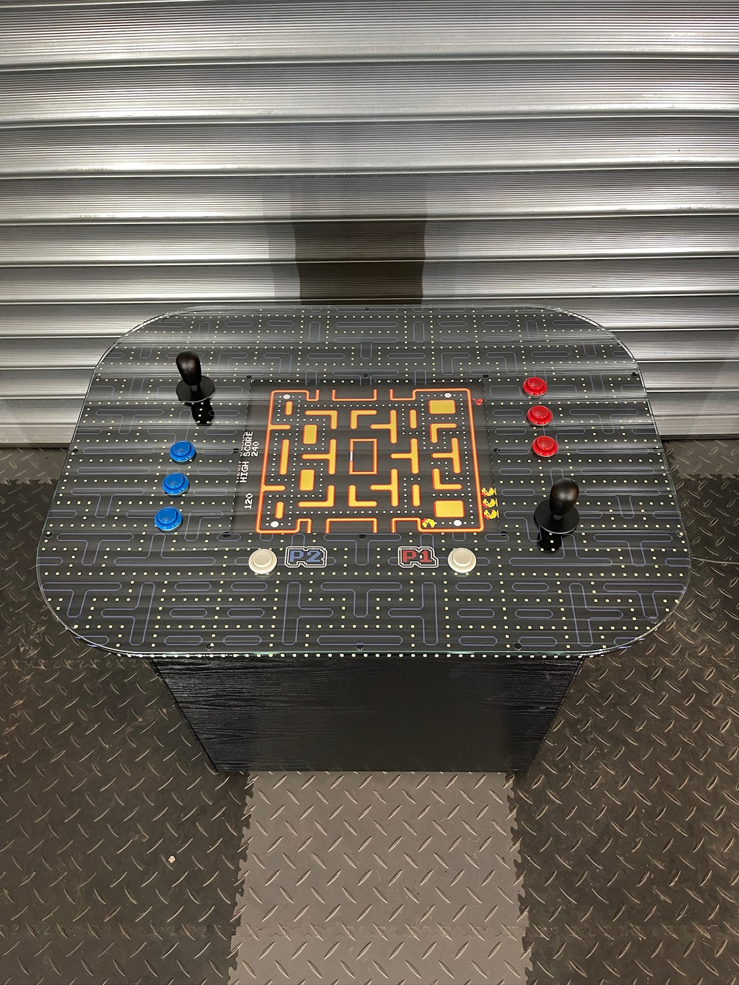 A retro arcade cocktail table with a glass top featuring colorful arcade game icons, built-in speakers, and control buttons on the edge.
