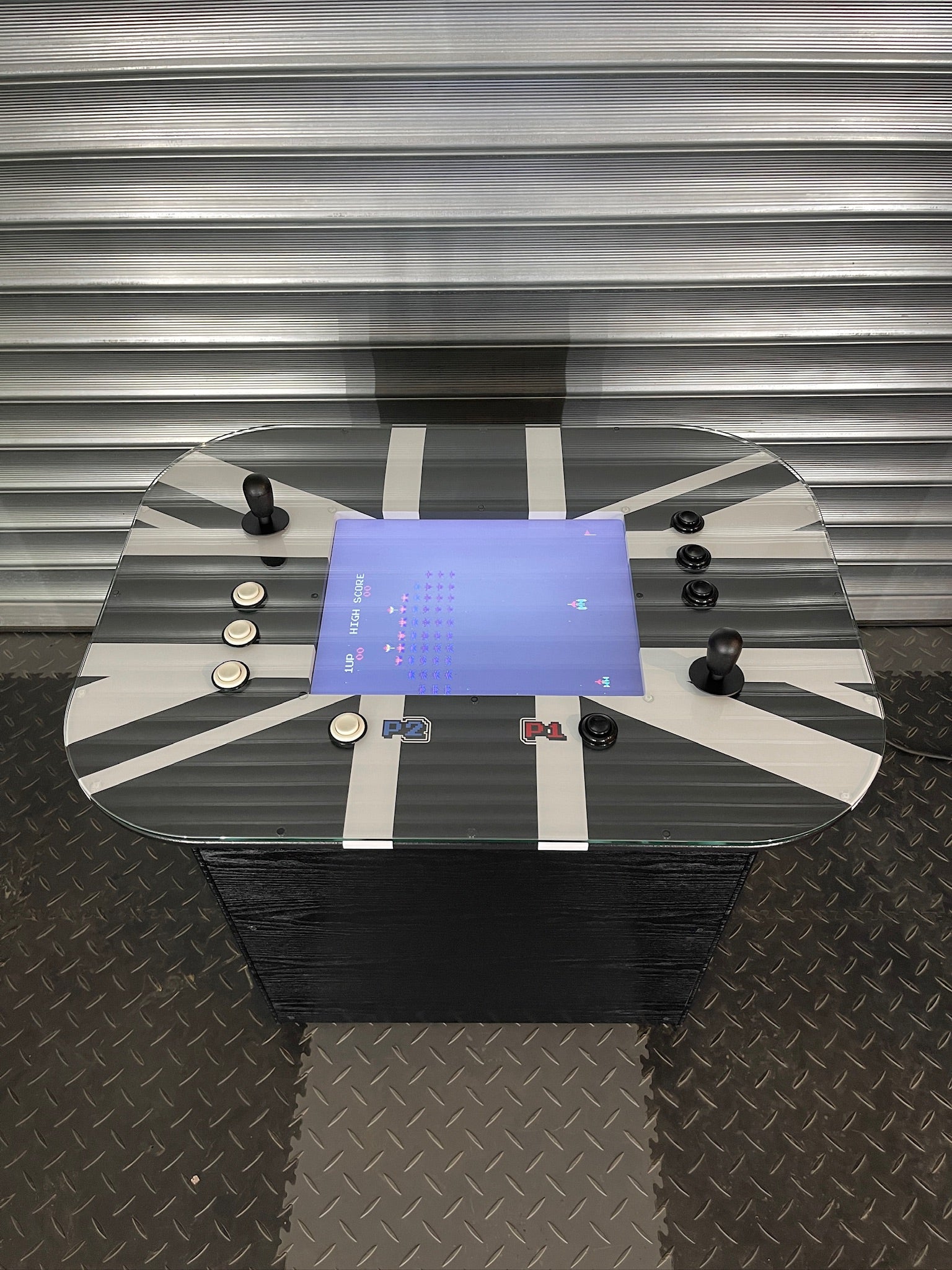 A retro arcade cocktail table with a glass top featuring colorful arcade game icons, built-in speakers, and control buttons on the edge.